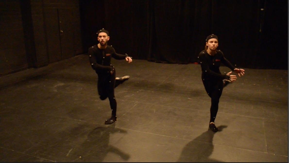 Two dancers in motion-capture suits with markers performing in a black studio