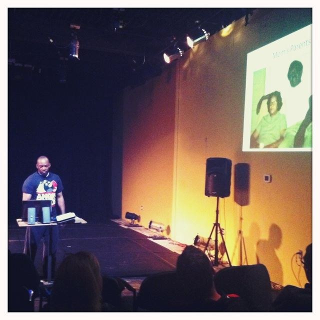 Boris Willis performing Black Merce on stage beside a projected image as the audience builds the score